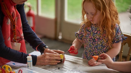 A child holding scissors doing a crafts stood beside a volunteer activity assistant